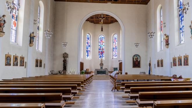 Kirche von Innen. Hell erleuchtet mit Blick auf den Rundbogen vorne am Altar. Links und rechts sind in Reihen Holzbänke.