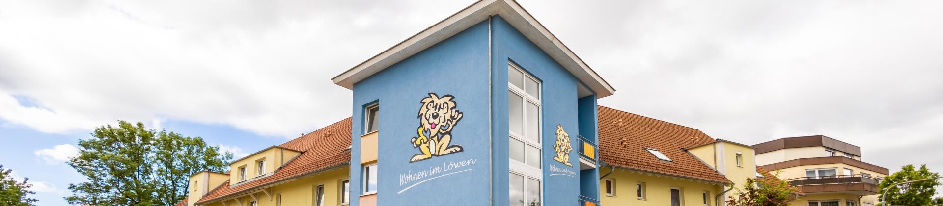 Wohnanlage Löwen von außen. Blau-gelbes Gebäude mit dem Logo der Wohnanlage dem "Löwen"