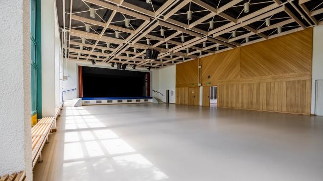 Die Festhalle von innen. Ein großer langer Raum, mit einer Bühne, vielen Lampen an der Decke und einer Wandverkleidung aus Holz an der rechten Seite.