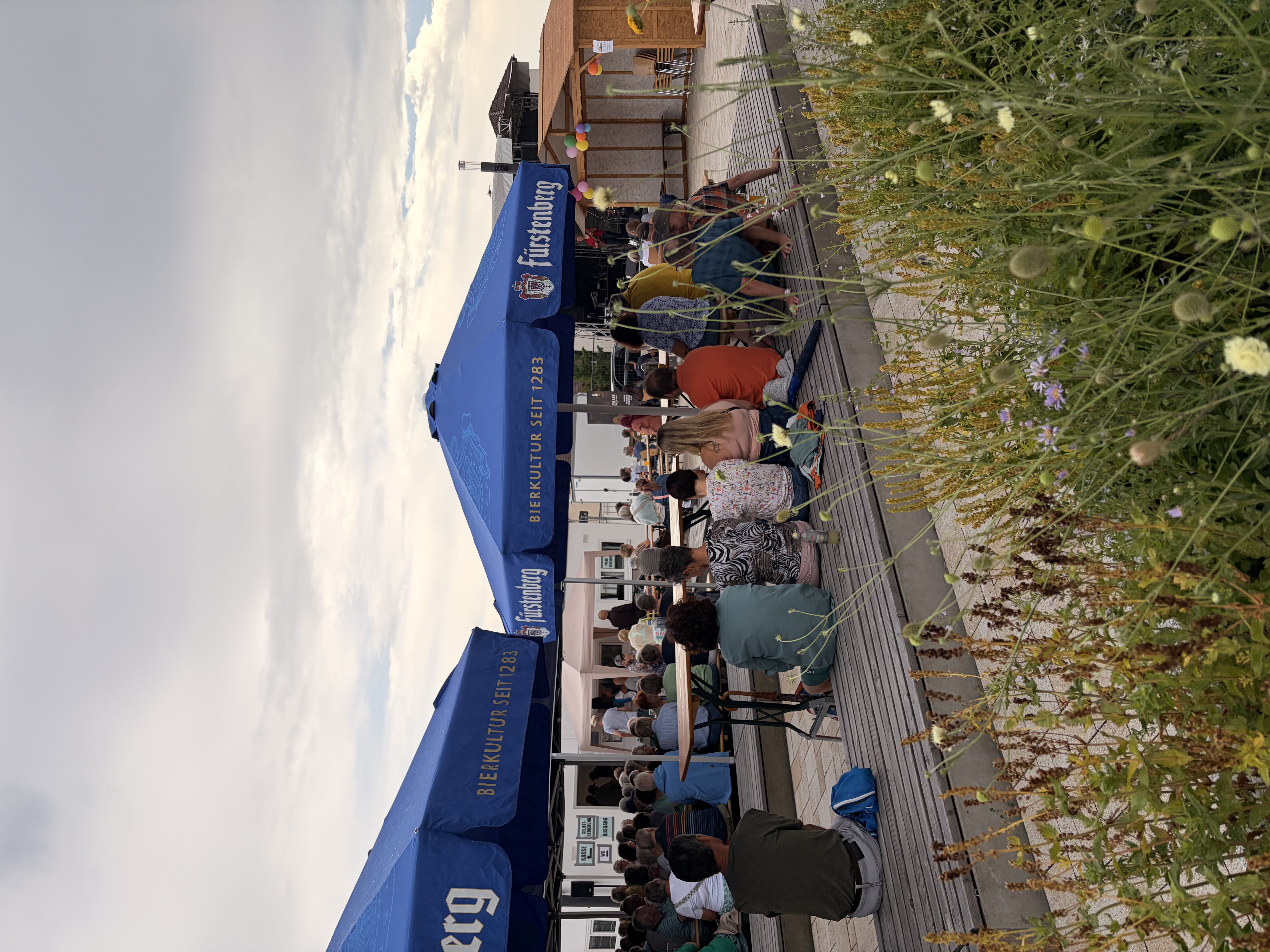 Leute flanieren auf dem Dorfplatz, der inzwischen schön grün bewachsen ist.