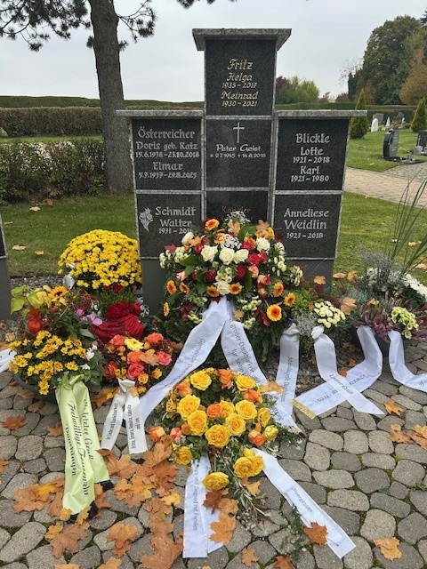 Viele bunte Trauerkränze mit Schleifen vor der Urnenwand in grau.