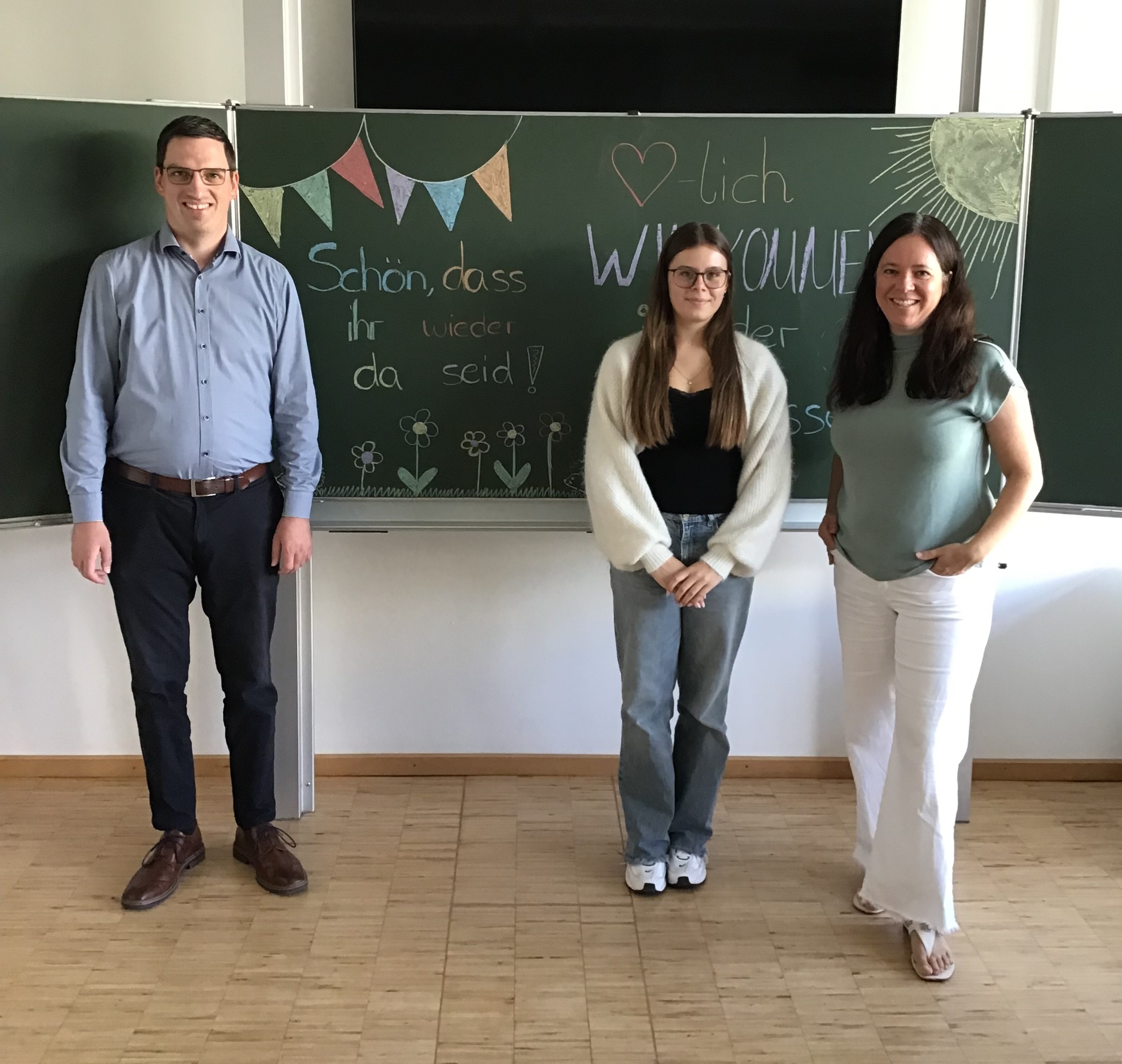 Hauptamtsleiter Andreas Krebs, FSJ-Kraft Lena Edeler und Rektorin Kathrin Michl stehen anlässlich der Begrüßung vor einer Tafel in einem Klassenzimmer, die bunt bemalt ist.