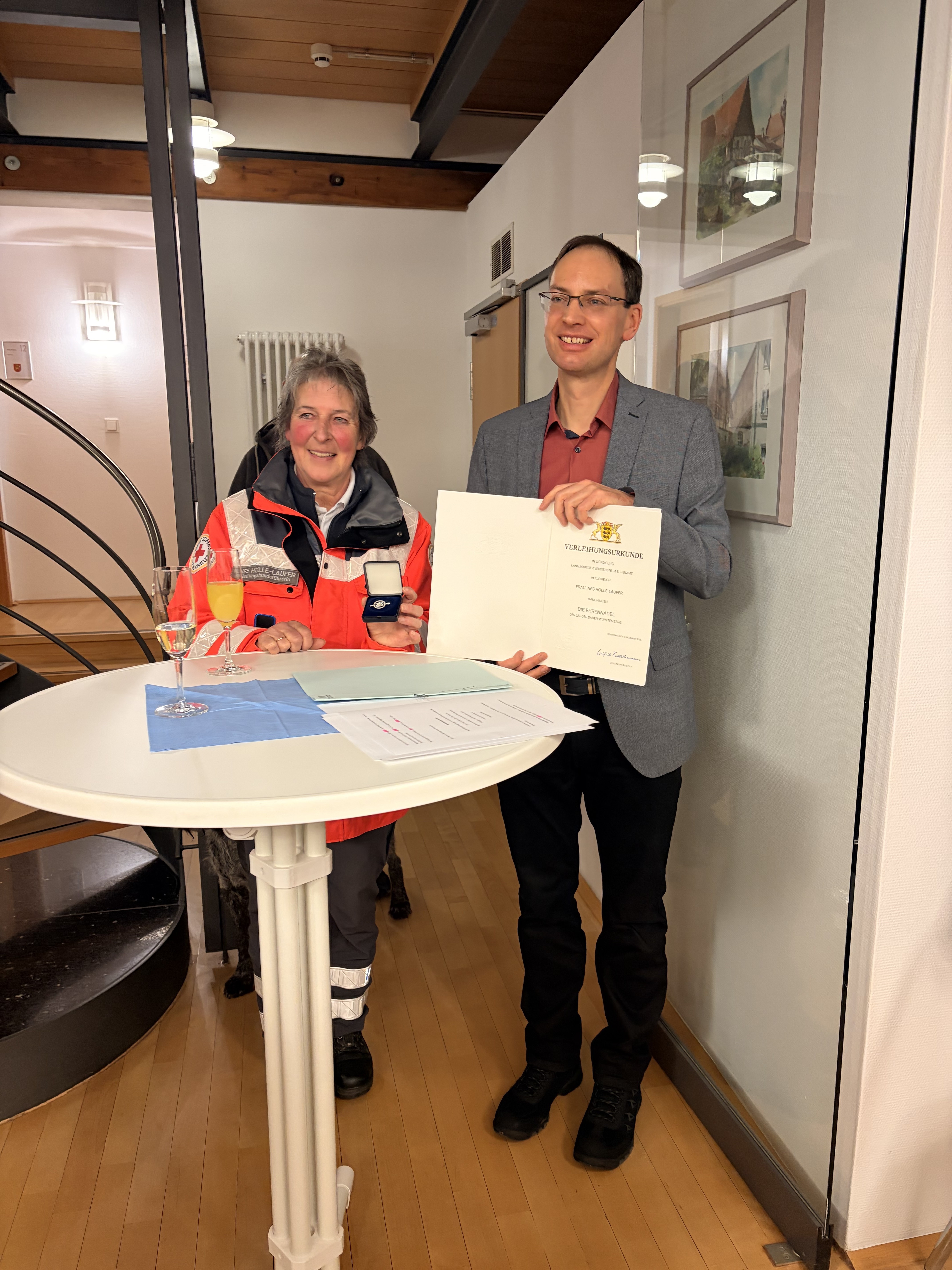 Links Frau Ines Hölle-Laufer vor einem Bistro-Stehtisch, rechts daneben Bürgermeister Torben Dorn mit der Urkunde in der Hand.